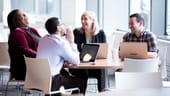 Photo of four colleagues sitting at a table with laptops open, laughing at a joke.
